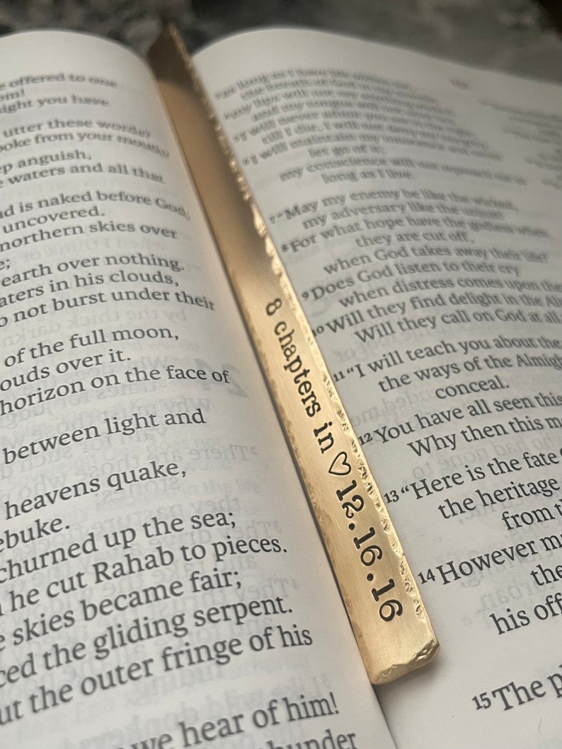 May include: A gold-colored bookmark with the text "8 chapters in 12.16.16" engraved on it. The bookmark is lying on top of an open book.