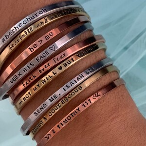 May include: A stack of silver, gold, and copper bracelets with inspirational messages engraved on them. The bracelets include phrases like "#bethechurch", "he > me", "SEEK HIS FACE", "faith over fear", "NOT MY WILL BUT YOURS", "SEND ME. ISAIAH 618", "strong & courageous", and ">>> 2 TIMOTHY 1:7 <<<<".
