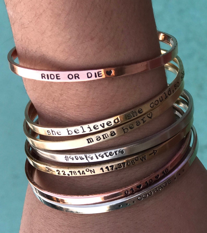 May include: A stack of gold and silver bracelets with engraved messages, including "Ride or Die", "She believed she could, so she", "Mama bear", "#soulsisters", "22.7814&deg;N 117.3768&deg;W", "01.10.16", and "...COURAGE...".