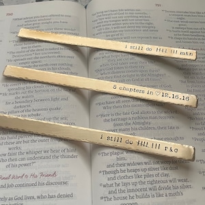 May include: Two wooden sticks with the text "i still do HHH III m&n" and "i still do HHH III r&c" written on them. The sticks are resting on an open book with text about the book of Job from the Bible.
