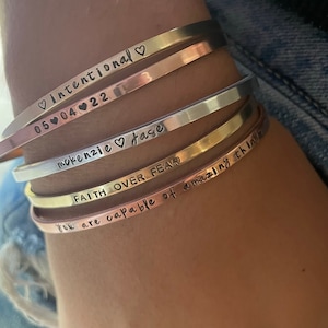 May include: A set of five gold, silver, and rose gold bangle bracelets with engraved inspirational messages. The messages include "intentional", "05-04-22", "mokenzie jase", "FAITH OVER FEAR", and "You are capable of amazing things".