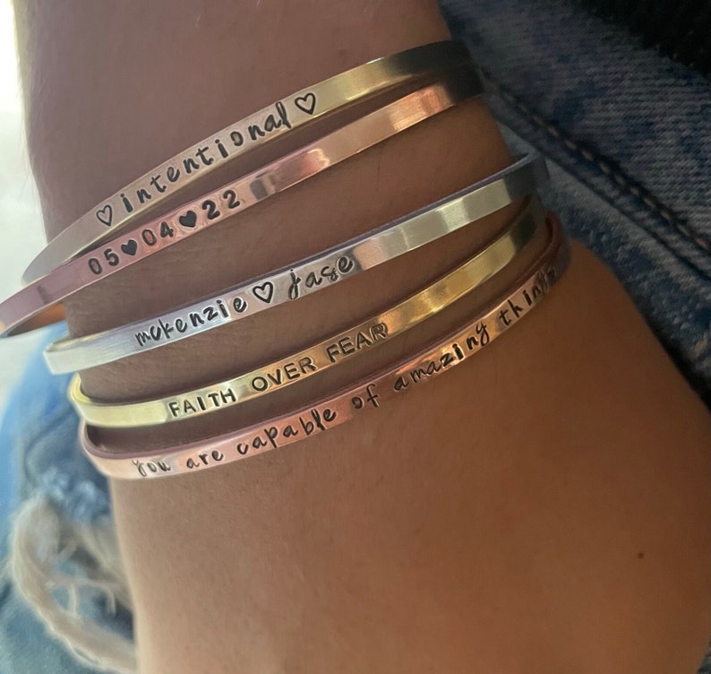 May include: A set of five gold, silver, and rose gold bangle bracelets with engraved inspirational messages. The messages include "intentional", "05-04-22", "mokenzie jase", "FAITH OVER FEAR", and "You are capable of amazing things".