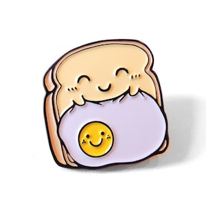Op de afbeelding: Een cartoon emaille pin van een glimlachende snee brood die een gebakken ei met een smiley vasthoudt. Het brood is lichtgeel en het ei is paars met een gele dooier.