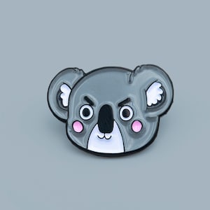 Può includere: Spilla in smalto a forma di koala grigio con contorno nero. Il koala ha un'espressione severa con guance rosa e dettagli bianchi. La spilla è posta su uno sfondo grigio.