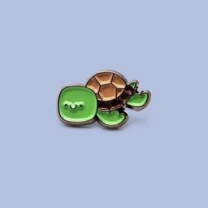 Puede incluir: Un pin de esmalte de tortuga verde y marrón. La tortuga tiene una cara verde cuadrada con una boca sonriente, un caparazón marrón y aletas verdes. El pin está sobre un fondo azul claro.