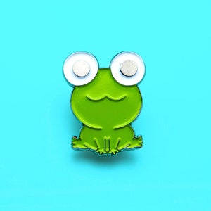 Op de afbeelding: Een groene kikker-vormige emaille pin met grote witte en zilveren ogen. De pin is geplaatst tegen een helderblauwe achtergrond. De kikker lacht en heeft een eenvoudig, cartoonachtig ontwerp.