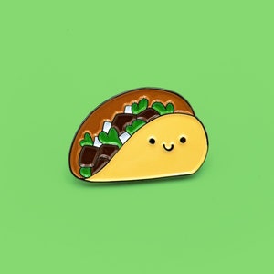 Può includere: Una simpatica spilla a forma di taco smaltato con un volto sorridente. La spilla presenta un guscio marrone, con ripieni di carne marrone, cipolle bianche e lattuga verde. Lo sfondo è verde chiaro.