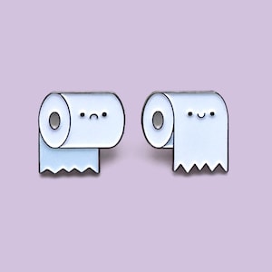 Op de afbeelding: Twee lichtblauwe toiletpapierrolspelden met zwarte contouren en lachende gezichten. Eén speld heeft een verdrietig gezicht, de andere een blij gezicht. De achtergrond is effen lavendel van kleur.