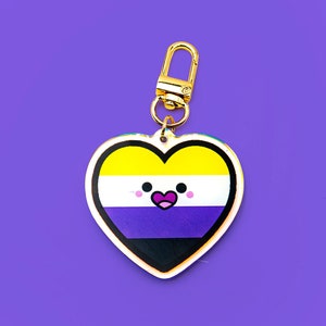 Op de afbeelding: Een hartvormige sleutelhanger met een goudkleurige sluiting. Het hart toont de kleuren van de non-binaire pride-vlag: geel, wit, paars en zwart, met een schattig gezichtsontwerp. De achtergrond is effen paars.