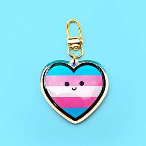 Op de afbeelding: Een gouden hartvormige sleutelhanger met een transgender pride vlag ontwerp. Het hart heeft een glimlachend gezicht.