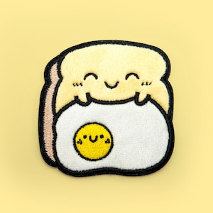 Op de afbeelding: Een gele geborduurde patch van een glimlachende cartoon broodschijf die een wit ei met een geel eigeel vasthoudt. Het ei heeft een glimlachend gezicht.