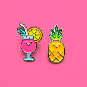 Op de afbeelding: Twee emaille pins op een roze achtergrond. Eén pin is een roze cocktail met een lachend gezicht, een rietje en een schijfje citroen. De andere is een gele ananas met een lachend gezicht en groene bladeren.