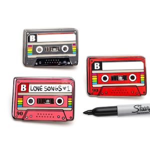 Könnte beinhalten: Drei Emaille-Pins in Form von Kassetten. Die Pins sind rot, schwarz und pink. Die pinkfarbene Kassette trägt den Text "B LOVE SONGS v1". Die Pins befinden sich auf einem weißen Hintergrund.
