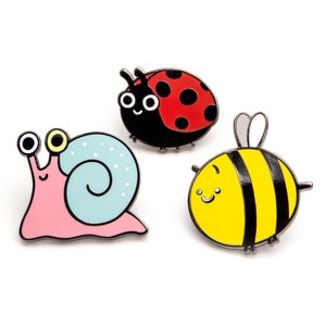Op de afbeelding: Drie emaillepins met een roze slak met een blauwe schelp, een rode lieveheersbeestje met zwarte stippen en een gele en zwarte bij. De pins zijn allemaal in cartoon-stijl en hebben glimlachende gezichten.