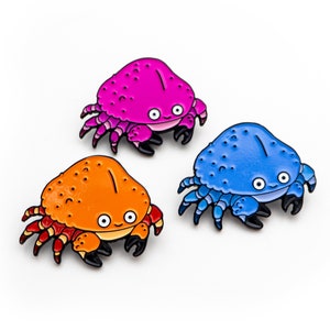 Op de afbeelding: Drie kleurrijke krab emaille pins. Eén is felroze, een andere is oranje en de derde is blauw. Elke pin heeft een lachend gezicht, zwarte klauwen en een witte achtergrond.