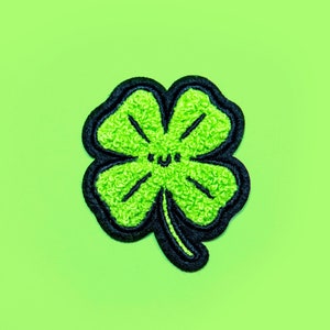 Op de afbeelding: Een groene klaverblad patch met een zwarte rand en een lachend gezicht. De klaver heeft een getextureerde, fuzzy uitstraling. De achtergrond is een effen, heldergroene kleur.