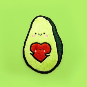 Op de afbeelding: Een groene avocado-vormige patch met een glimlachend gezicht en een rood hart met een glimlachend gezicht erin. De patch is gemaakt van vilt en heeft een gestikte omtrek.