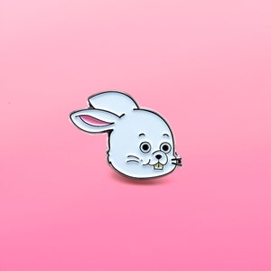 Op de afbeelding: Witte emaille pin van een cartoon konijn met een brede glimlach en grote ogen. Het konijn heeft een roze neus en een witte achtergrond.