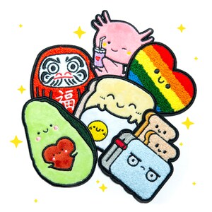 May include: A set of nine embroidered iron-on patches featuring various cute characters and designs, including a red and white daruma doll, a rainbow heart, a smiling avocado, a blue toaster, a yellow egg, and a pink axolotl.