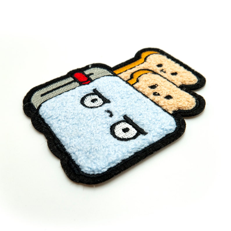May include: A blue and black embroidered patch of a cartoon toaster with a grumpy expression and three slices of bread inside. The bread is embroidered in a light brown color.