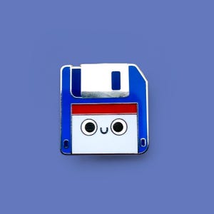 Op de afbeelding: Een blauw, wit en rode emaille pin in de vorm van een floppy disk met een lachend gezicht. De pin heeft een zilveren rand en staat tegen een blauwe achtergrond. Het ontwerp is een retro, nostalgische knipoog naar vintage technologie.