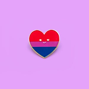 Puede incluir: Un pin en forma de corazón rojo, rosa y azul con una cara sonriente. El pin representa la bandera del orgullo bisexual.