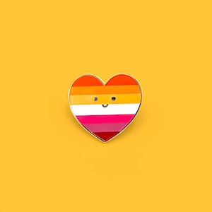 Op de afbeelding: Een hartvormige emaille pin met een gouden rand, met de kleuren van de lesbische pride-vlag: oranje, roze tinten en wit. De pin heeft een lachend gezicht. De achtergrond is effen geel.