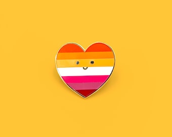 May include: A heart-shaped enamel pin with a gold border, featuring the lesbian pride flag colors: orange, shades of pink, and white. The pin has a smiling face design. The background is a solid yellow.
