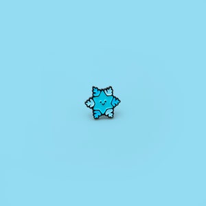 Op de afbeelding: Een blauwe emaille pin in de vorm van een sneeuwvlok met een glimlachend gezicht. De pin heeft een zwarte omtrek.