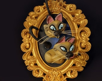 May include: A whimsical, golden-framed artwork featuring two cartoon cats with tan fur, pink ears, and blue eyes. The cats are set against a dark blue background, with one cat's tail arching above the frame. The frame has ornate detailing.