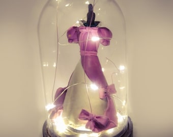 May include: A glass dome with a small purple and white dress figurine inside. The dome is lit with white fairy lights.