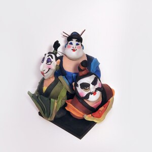May include: A hand-painted figurine set of three figures with exaggerated features. The figures are wearing traditional Chinese clothing and have colorful faces. The figurine set is on a black base.