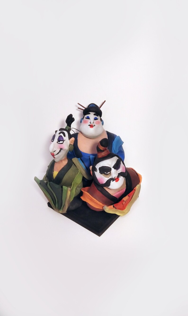 May include: A hand-painted figurine set of three figures with exaggerated features. The figures are wearing traditional Chinese clothing and have colorful faces. The figurine set is on a black base.