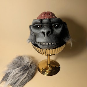 May include: A quirky sculpture of a gorilla's head with a visible brain, displayed on a gold-coloured pedestal. The gorilla's face is grey with detailed features, and a piece of grey fur is next to the pedestal.