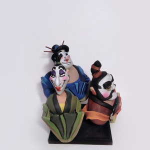 May include: Three hand-painted ceramic figurines of people wearing traditional Japanese clothing. The figurines are painted in black, white, blue, green, yellow, orange, and red. The figurines are standing on a black base.