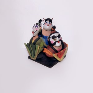 May include: A hand-painted ceramic figurine of three people in traditional Japanese clothing. The figurine is painted in bright colors, including red, blue, green, and yellow. The people are smiling and have exaggerated features.