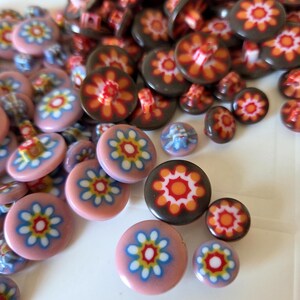 May include: A collection of round buttons in various sizes, featuring a central floral pattern with white, yellow, and blue petals on a pink base, or orange, yellow, and white petals on a dark brown base. Each button has a shank on the back.
