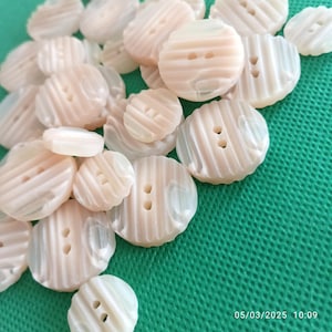 Pode incluir: Uma coleção de botões brancos e creme com um design texturizado e nervurado. Os botões têm dois furos e estão dispostos em uma pilha sobre uma superfície verde.