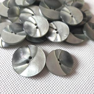 Op de afbeelding: Een verzameling parelmoer grijze knopen met een tweekleurig ontwerp. Elke knoop heeft twee gaten en een wervelpatroon. De knopen zijn rond en lijken van plastic te zijn.
