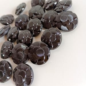 Puede incluir: Una colección de botones redondos de vidrio negro con un diseño floral en relieve. Los botones están dispuestos en semicírculo sobre una superficie blanca.