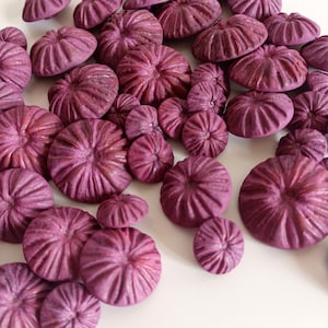 May include: A collection of purple, round, decorative buttons with a raised, textured design. The buttons are arranged in a random pattern on a white surface.