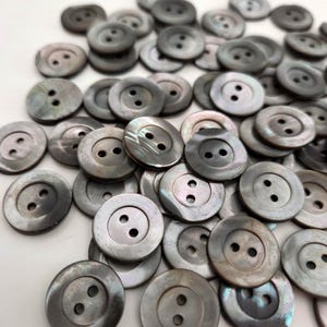 Puede incluir: Un conjunto de botones redondos grises, cada uno con dos agujeros. Los botones tienen un brillo nacarado y están dispersos sobre una superficie blanca. Adecuados para costura y proyectos de manualidades.