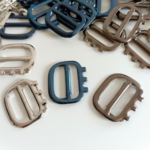 May include: Assortment of metal slider buckles in silver, gold, and blue. The buckles are rectangular with a single opening and a series of small teeth on the side.