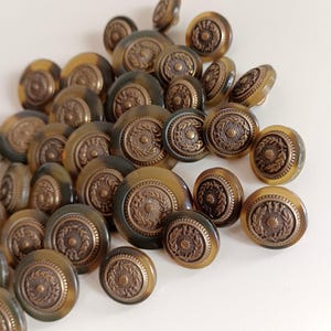 Könnte beinhalten: Ein Haufen brauner und goldener Knöpfe mit einem dekorativen Design. Die Knöpfe sind rund und haben ein erhabenes Design aus einem Kranz mit einem kleinen Kreis in der Mitte.