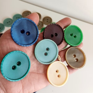 Peut inclure: Assortiment de boutons colorés avec deux trous au centre. Les boutons sont en plastique et ont une finition lisse et brillante. Les couleurs incluent le bleu, le vert, le marron, le crème et le rouge.