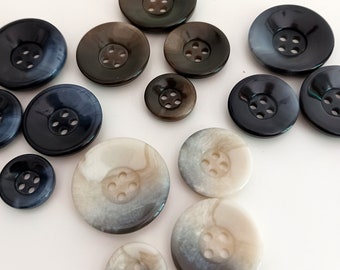 Könnte beinhalten: Sortiment von 12 Knöpfen in verschiedenen Farben und Mustern, darunter Schwarz, Braun, Weiß und Grau. Die Knöpfe haben jeweils vier Löcher und sind aus einem glatten, glänzenden Material gefertigt.