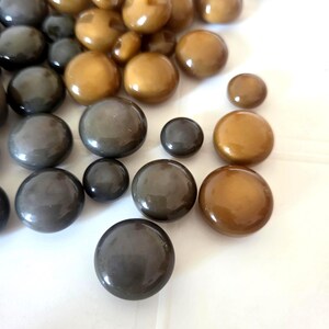 May include: A selection of glossy, round buttons in various sizes and shades of brown and grey. The buttons have a smooth, reflective surface, and are scattered across a white surface. The buttons appear to be made of a hard, durable material.