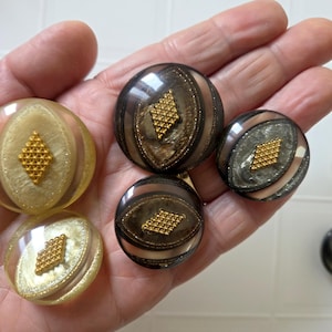 May include: A collection of five large, decorative buttons. The buttons feature a clear, domed design with a gold diamond-shaped embellishment in the center. The buttons have a variety of colors, including gold, brown, and silver.