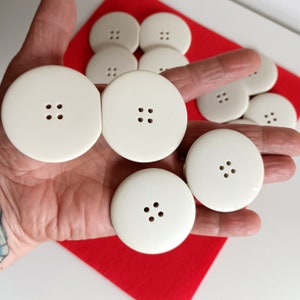 Puede incluir: Una colección de diez botones de plástico blanco con cuatro agujeros cada uno. Los botones están dispuestos en círculo sobre una superficie roja.