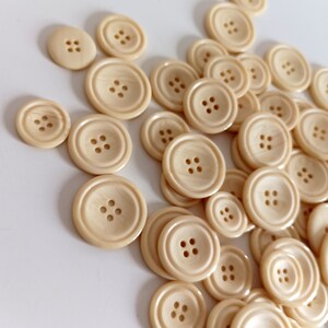 May include: A collection of off-white plastic buttons of various sizes. The buttons have four holes each and are arranged in a random pattern.